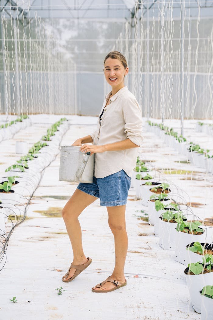 Smiling woman in greenhouse holding a watering can, tending to potted plants.